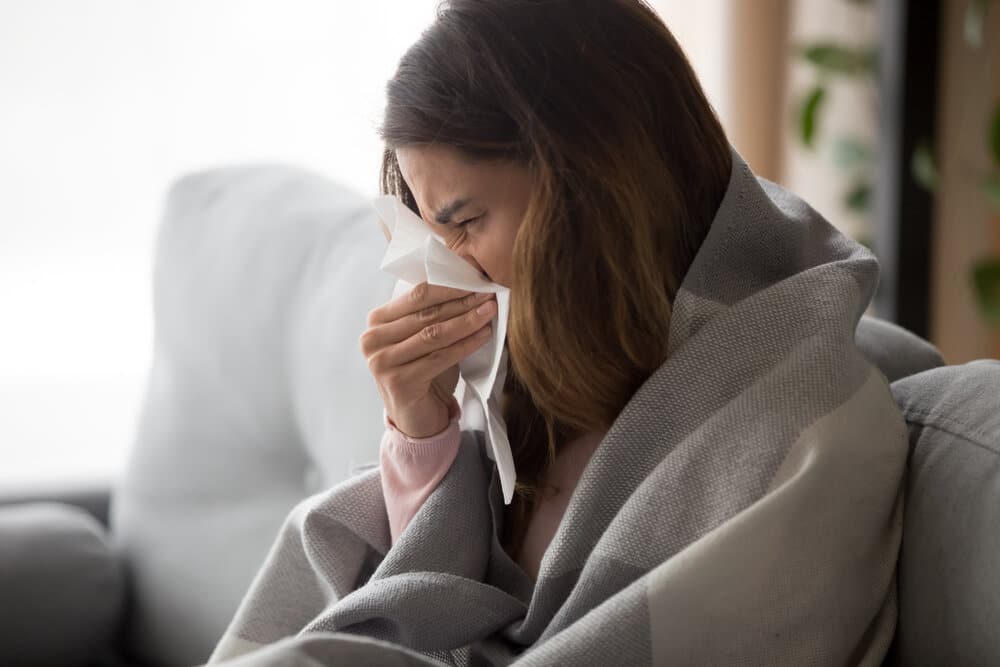 Ill upset young woman sitting on sofa covered with blanket freezing blowing running nose got fever caught cold sneezing in tissue, sick girl having influenza symptoms coughing at home, flu concept